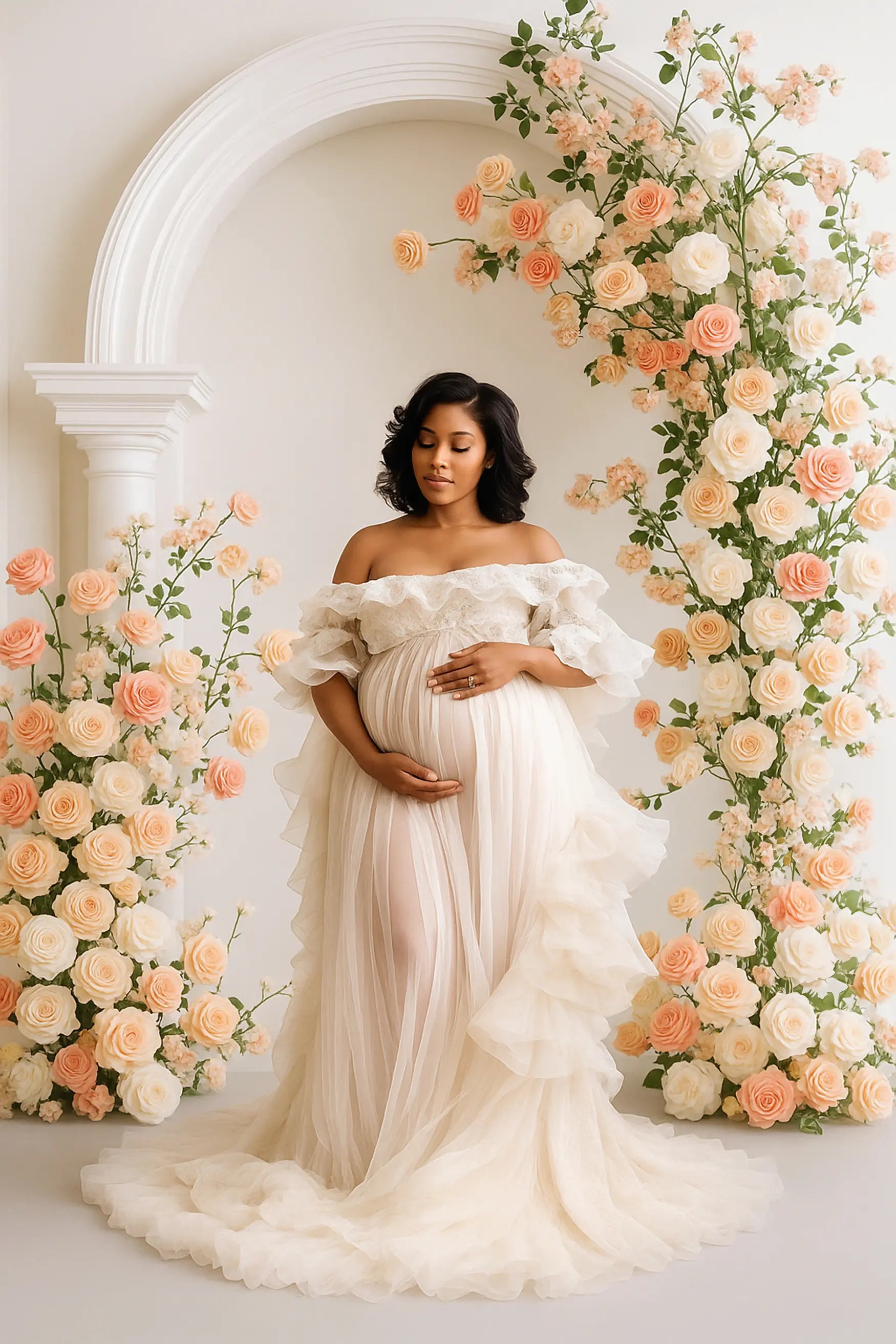 Pregnant woman in a white gown standing between floral arches with peach and white flowers.